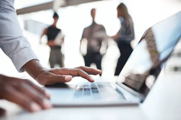 Close-up of a person using a laptop with blurred colleagues in the background.